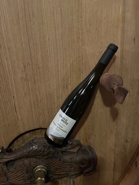 Bouteille de Pinot Gris Grand Cru Frankstein 2023 appuyée contre le fût de chêne de 1000 L qui l’a élevé ; capsule noire mate, lattes patinées du tonneau, lumière rasante qui révèle les reflets dorés du vin