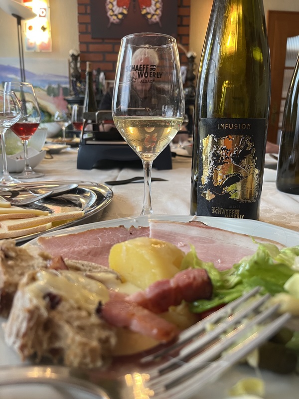 Bouteille de vin Auxerrois Infusion 2024 d'Alsace (avec infusion de Gewurztraminer) servie avec un plat de raclette et de la charcuterie.